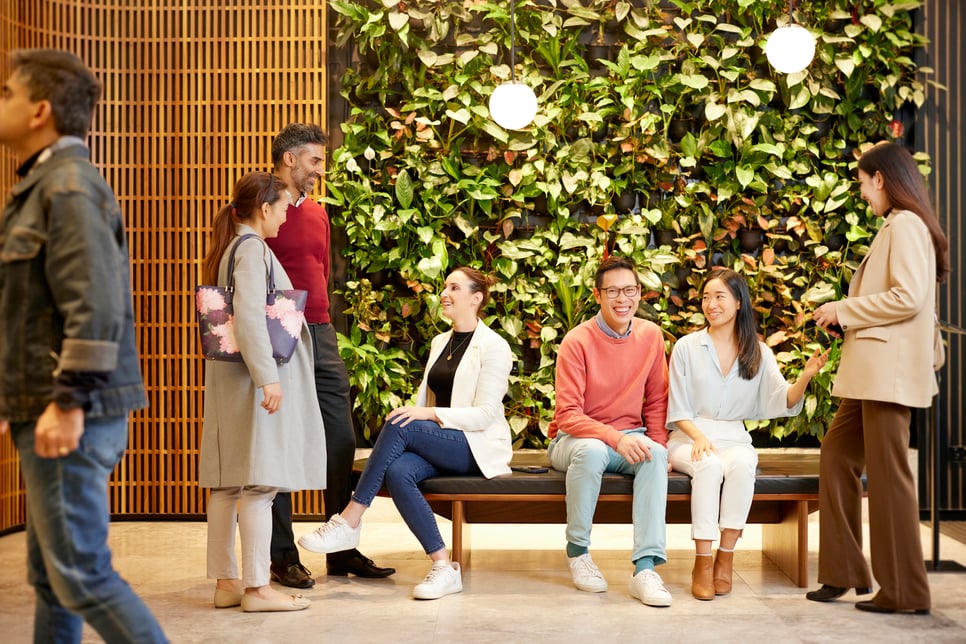 A group of individuals smiling and talking, sitting on a bench in front of greenery